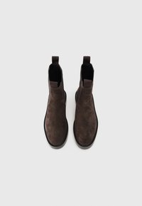 Une paire de bottes Chelsea en daim brun foncé avec des panneaux élastiques noirs sur les côtés et des tirants, photographiée de dessus sur un fond blanc.