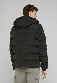 Bărbat purtând o geacă neagră căptușită cu glugă și pantaloni cargo gri deschis, cu spatele spre cameră, pe un fundal simplu gri deschis.