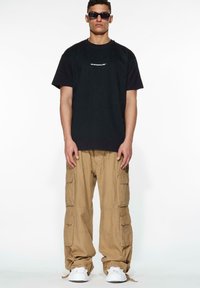Black cotton T-shirt with white text, paired with beige cargo pants featuring multiple pockets and a loose fit. Worn with white sneakers.