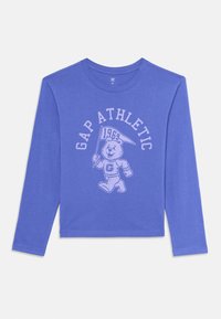Kék hosszú ujjú ing, amelyen a "GAP ATHLETIC" felirat és egy medvemaszkot tartó zászlót lengető medve látható, amin a "1969" felirat áll.