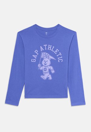 Chemise à manches longues bleue avec le texte "GAP ATHLETIC" et une mascotte d'ours tenant un drapeau portant "1969" sur le devant.
