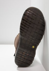 Calzado marrón con una suela de goma texturizada, que presenta un patrón de tacos y un pequeño emblema amarillo. El diseño incluye ojales metálicos para los cordones.
