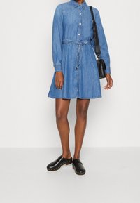 Farkkumekko, jossa pitkät hihat, napit edessä, vyötärönauha ja levenevä helma. Yhdistetty mustiin pukuihin, joissa koristeelliset niitit.