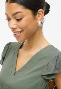 Blusa de manga corta color verde oliva con escote en V y volantes en las mangas. La modelo lleva pendientes de gota en plata, mostrando una textura suave.