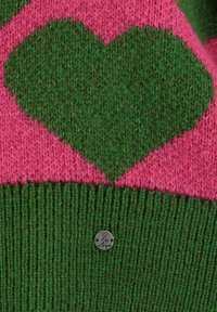 Motivo a forma di cuore verde su tessuto di maglia rosa sopra un maglione verde a coste con un piccolo cartellino metallico rotondo attaccato.