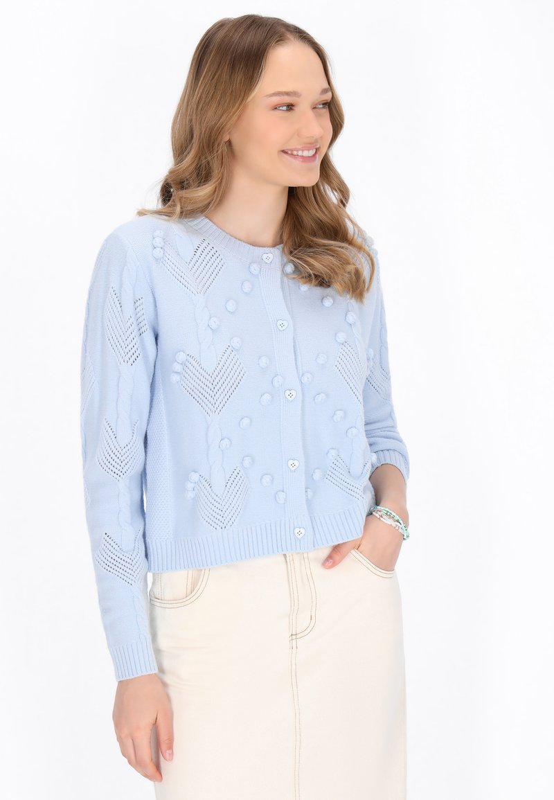 Vaalea sininen neulottu neuletakki, jossa on tekstuurikuvioita, pyöreä pääntie, nappikiinnitys ja resorikäänteet. Yhdistetty beigeen hameeseen.