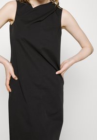 Černé šaty bez rukávů s přiléhavým střihem a splývavým výstřihem. Vyrobeno z hladkého materiálu, bez viditelných vzorů nebo akcentů.