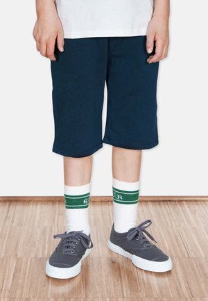 Marineblaue Shorts mit lockerer Passform, kombiniert mit weißen Socken mit grünen Streifen und grauen Sneakers mit weißen Sohlen.