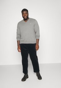 Grijze V-halstrui van zachte stof, gedragen met donkerblauwe corduroy broek en zwarte enkellaarsjes. Model staat tegen een effen achtergrond.