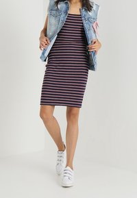 Femme portant une robe sans manches rayée bleu marine, un gilet en denim bleu clair avec un ruban rose, et des baskets blanches à velcro marchant en avant.