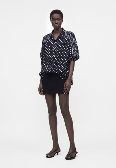 Γυναίκα με μαύρο και λευκό πουκάμισο με κουμπιά και φουσκωτά μανίκια, μαύρη μίνι φούστα και μαύρα σανδάλια με χαμηλό τακούνι, στέκεται ακίνητη.