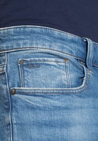 Vaaleansiniset farkut, joissa on hieman kulunut ilme, etutasku, jossa tummat metalliset niitit ja tikattu "RAW"-logodetalji.