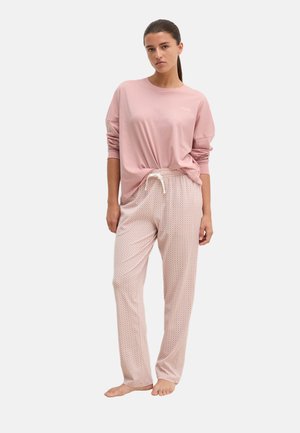 Vrouw staat blootsvoets, draagt een roze shirt met lange mouwen en lichtroze pyjamabroek met patroon en een trekkoord in de taille.