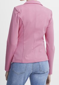 Pink blazer with a structured design, featuring long sleeves, a notched collar, and smooth fabric. Paired with light blue denim jeans.