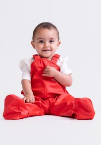 Helle rote wasserdichte Overalls mit verstellbaren Trägern, die eine glatte, glänzende Textur aufweisen. Kind sitzt, Hände gefaltet, lächelt.