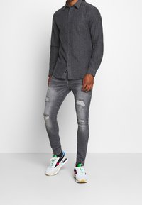 Man in een donkergrijs overhemd met knopen, versleten skinny grijze spijkerbroek en witte sneakers met zwarte, groene, rode en blauwe details.