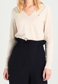 Beżowy sweter w serek z miękkiego materiału, połączony z wysokim stanem, ciemnonawigacyjnymi spodniami z marszczoną talią i bocznymi kieszeniami.