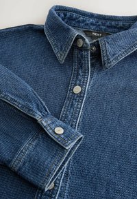 Tumedast sinises denimpajamas on tekstuuriga muster. Omab kraega kaelust, nööbistust ja sobiva materjaliga nööbitavaid varrukate otsi.