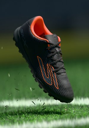 Chaussure de football noire avec intérieur et design orange, suspendue au-dessus d'un terrain herbeux avec des brins d'herbe volant autour.