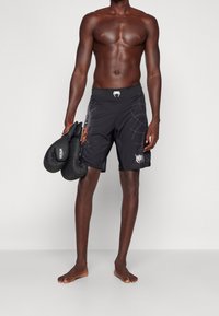 Shorts de boxeo negros con detalles gráficos y logo, combinados con guantes de boxeo negros, usados por una persona de pie descalza frente a un fondo neutral.