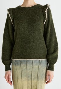 Pull vert foncé en tissu duveteux avec des détails volantés aux épaules et une bordure beige clair, associé à une jupe en jean délavé taille haute avec des coutures verticales.