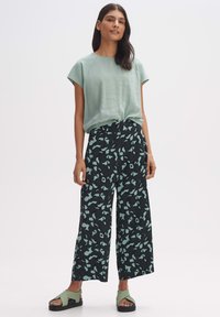 Lichtgroene top met korte mouwen, gecombineerd met een zwarte broek met wijde pijpen en een mintgroen abstract patroon. Het model draagt groene sandalen.