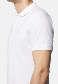 Biała koszulka polo z płaskim kołnierzykiem, krótkimi rękawami i małym niebieskim logo na lewej piersi, wykonana z fakturowanej tkaniny.