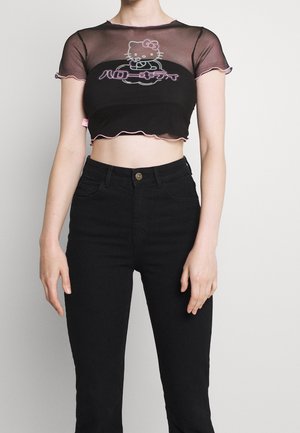 Zwarte mesh crop top met roze gerimpelde randen, voorzien van een Hello Kitty-graphic en Japanse tekst in een gedurfde vormgeving, gecombineerd met zwarte jeans.