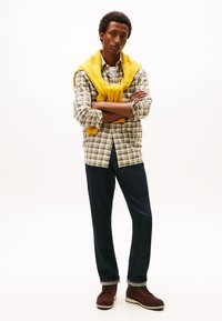 Camicia a quadri verde, beige e marrone; maglione giallo a maglia annodato attorno al collo; jeans blu scuro; stivali marroni con suole chiare.