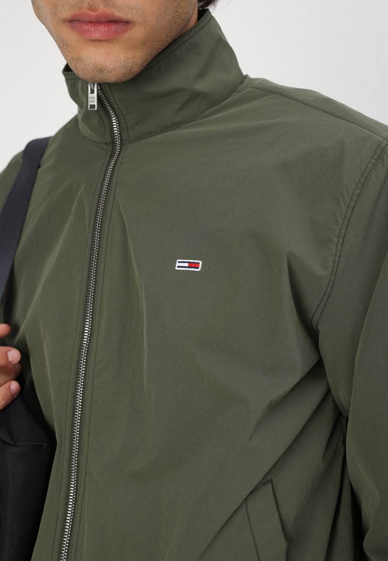 Veste zippée vert olive avec col montant et texture légère. Présente un petit patch logo en rouge, blanc et bleu sur la poitrine.