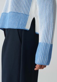 Person som bär en ribbad stickad tröja med blå och vita vertikala ränder och en sidoklyvning, tillsammans med mörkblå byxor.