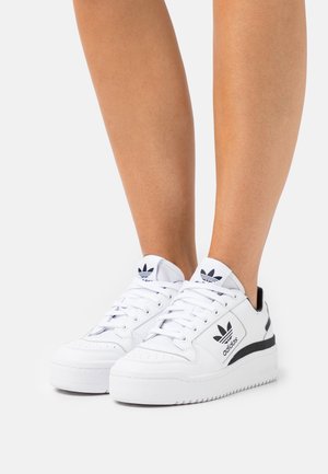 Baskets blanches Adidas avec logo noir et accents, portées sur jambes nues, inclinées pour montrer les vues latérale et avant.