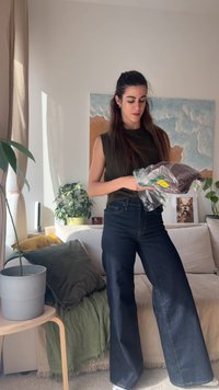 Žena stojí v obývacím pokoji a drží složené oblečení zabalené v plastu, obklopena pokojovými rostlinami a polštáři na pohovce.