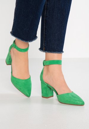 Pieds portant des chaussures à talons carrés en daim vert avec des brides à la cheville, associées à un jean bleu foncé à ourlet effiloché, posés sur un sol blanc.