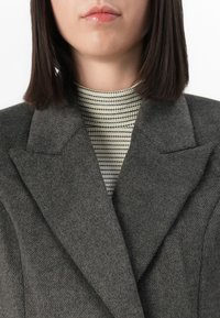 Femme portant un manteau en laine gris sur un col roulé crème et noir à rayures, avec des cheveux bruns foncés lisses jusqu'aux épaules.