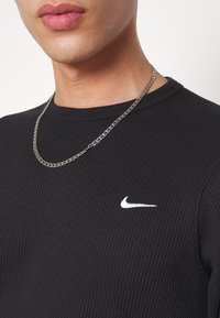 Chemise noire à manches longues à côtes, arborant un logo Nike blanc sur la poitrine, associée à un collier en chaîne argentée. Texture lisse et coupe ajustée.