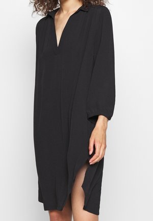 Zwarte, losvallende jurk met een kraag, diepe V-hals en lange mouwen. Heeft zijsplitten voor extra bewegingsvrijheid en een gladde textuur.