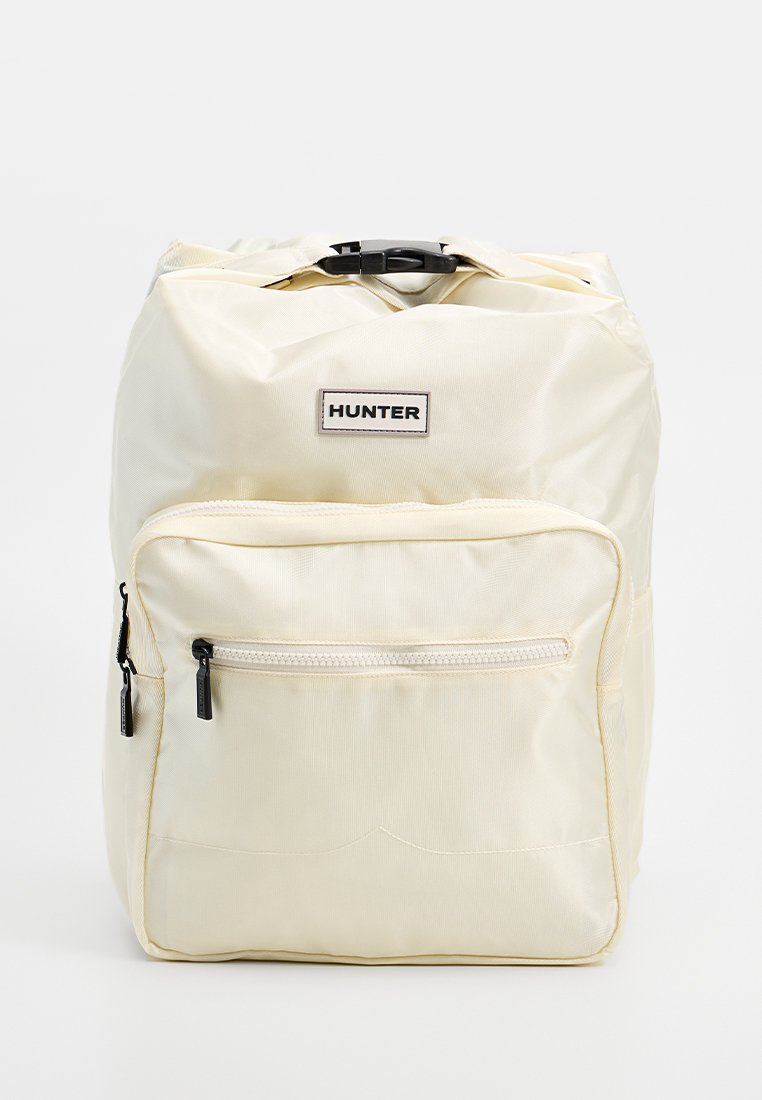 Mochila de color crema hecha de material brillante, con un bolsillo frontal con cremallera, correas ajustables y una etiqueta rectangular de marca.