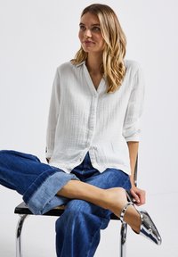 Ung kvinna som sitter med benen i kors på en stol, iförd en vit skjorta med knappar, uppvikta blå jeans och glittrande silverfärgade ankelremsskor.