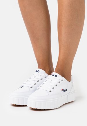 Sneakers platform bianche con design con lacci, caratterizzate dal logo Fila in blu e rosso sul lato. Materiale sintetico liscio, suola testurizzata.