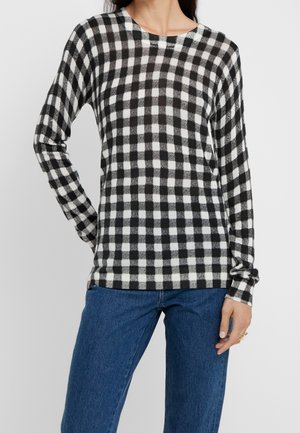 Kvinde iført sort- og hvidternet langærmet skjorte kombineret med blå jeans, stående foran en ensfarvet lys baggrund.