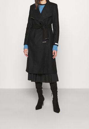 Femme portant un long manteau noir ceinturé sur un pull bleu, une jupe noire et des bottes noires à talons montant jusqu'aux genoux, debout devant un fond uni.