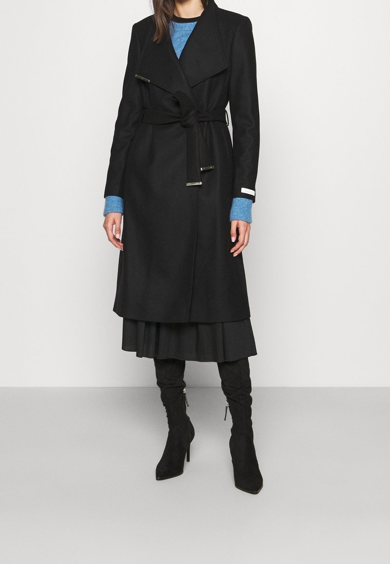 Femme portant un long manteau noir ceinturé sur un pull bleu, une jupe noire et des bottes noires à talons montant jusqu'aux genoux, debout devant un fond uni.