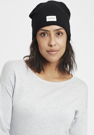 Black ribbed knit beanie with a white rectangular logo patch. The model wears a light gray, long-sleeve shirt with a wide neckline.