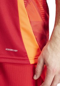 Maglietta sportiva rossa con accenti arancioni. Presenta un motivo testurizzato e il logo "AEROREADY". Realizzata in materiale traspirante.