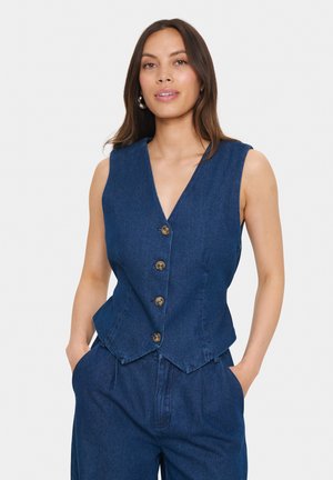Dunkelblaues Denim-Westen mit V-Ausschnitt, fünf braunen Knöpfen und einem ausgestellten Saum. Kombiniert mit dunkelblauen Hosen, die seitliche Taschen haben.