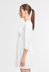Witte jurk met driekwart mouwen, een gerimpelde taille en een vloeiende rok, voorzien van een gestructureerde stof en een ronde halsontwerp.