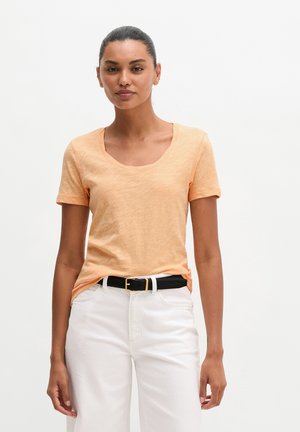 Kobieta o ciemnych włosach związanych do tyłu, ubrana w jasnopomarańczową koszulę z krótkim rękawem wpuszczoną w białe spodnie z czarnym paskiem, stojąca na białym tle.