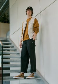 Mladý člověk v béžové vestě, hnědém svetru, černých kalhotách Nike, světlých vysokých teniskách a černé čepici stojící na vnitřním schodišti.