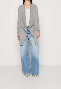 Light grey, open-front cardigan with long sleeves, worn over a beige top and light blue wide-leg jeans. Notable snake-print heels.